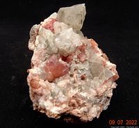 Photo of a pink amethyst mineral specimen displaying translucent pale pink crystals on a matrix with white and reddish mineral deposits, against a black background.
