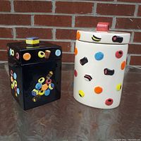 Photo showing two cookie jars, one black square and one white rounded rectangular, both decorated with colorful licorice allsorts candy designs on surface and lids.