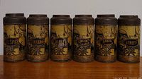 Front view of the 6 antique spice tins arranged side by side showing the labels Allspice, Cinnamon, Cloves, Cream of Tartar, Baking Powder, Ginger with visible age wear on metal lids and body.