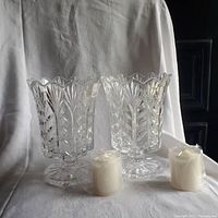 Photo showing a pair of clear crystal hurricane candle holders with intricate leaf pattern and scalloped rims, each paired with a white wrapped pillar candle in front.
