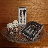 Glass tray, salt and pepper shakers, cheese spreaders in boxes shown on wood surface