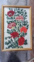 Full view of framed floral needlework featuring red and pink flowers with green leaves in a gold wood frame.