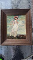 Front view of a wood framed print showing a young woman in white holding a parasol by the seaside under cloudy skies.