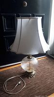 Full view of vintage table lamp with ribbed glass base, ornate metal cherub base, off-white fabric shade, and power cord with plug.