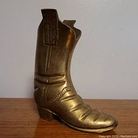 Side view of brass boot-shaped vessel showing detailed stitching and folds, heel, and flared opening