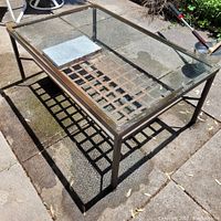 Top view of the rectangular metal garden table with glass top showing a lattice metal shelf underneath on a concrete patio.