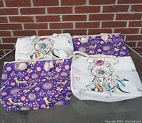 Four tote bags arranged on a surface showing two white bags with dreamcatcher design and two purple bags with woodland design and dreamcatchers, all with thick white rope handles.