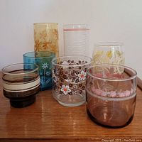 Seven vintage floral drinking glasses of varied colors and patterns on wooden surface.