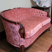 Front angle of vintage pink upholstered settee with carved wood arms and frame, showing rolled armrest and curved back.