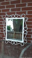 Full view of the white metal framed mirror with scrollwork against a brick wall and outdoor background.