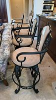 Four upholstered swivel bar stools with black metal scrollwork frames and tan fabric seats, arranged in a line indoors next to a kitchen counter and a sofa.