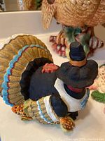 Close-up side view of a resin turkey pilgrim figurine wearing a black dress with white apron and black pilgrim hat, showing detailed feathers and corn decoration.