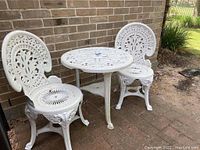Full view of the set showing two white cast aluminum chairs with intricate leaf and scroll designs and matching round table with patterned top beside a brick wall.