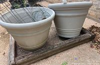 Two large beige plastic planters outdoors with two tomato cages inside, showing dirt and weathering.