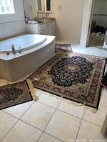 Area rugs in bathroom with large rug approximately 7x5 feet and two smaller rugs 2x4 feet. Black with gold and burgundy floral medallion pattern. Rugs have fringed edges and lie on tile floor near bathtub and sink.