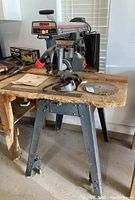 Radial saw with stand, blades, safety glasses, manual, ruler and wrenches on wooden table.
