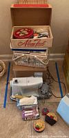 Box of sewing notions including buttons, pincushions, and small sewing accessories along with Singer sewing machine and steam iron in background