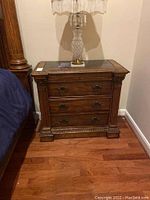 Wood nightstand with ornate carved detailing and three drawers, shown in full view with lamp on top.