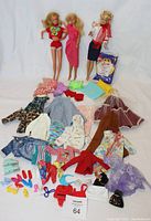Full shot of three Barbie dolls with various vintage doll clothes, shoes, and accessories spread out in front. One Barbie is in red shorts and a colorful top, another in a pink sheath dress, and a third in a pink jacket and skirt. Sealed McDonald's Barbie Rapunzel wedding doll in original packaging is visible.