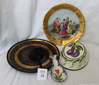 Photo showing four decorative hanging items: a gold-trimmed Limoges plate with a classical scene, a black metal candle holder plate with a central candle holder, a Delft-style ceramic spoon holder with floral and tulip design, and an Italian ceramic eggplant hanging piece.