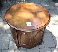 Photo showing round wooden table with sculpted edges, double doors with metal handles below, standing on stone patio.