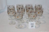 Photo showing eleven vintage pine cone-patterned wine glasses with gold detailing arranged in rows on white background.