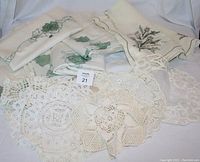 Five green cutout panel curtains with leaf designs, lying folded on a white fabric surface, alongside crocheted doilies and an embroidered tablecloth showing overall lot contents.