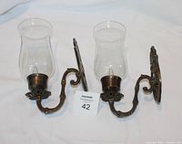 Pair of metal wall sconces with clear glass hurricane globes lying side by side on a white surface.