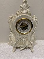 Full front view of the white porcelain ornate clock with baroque styling and gold-tone clock bezel.