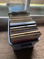Box of approximately 75 to 100 vinyl records stacked vertically inside a large plastic bin, seen in natural daylight.