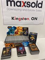 The lot shows a box filled with a large stack of World of Warcraft trading cards alongside several booster packs in colorful packaging, all set in front of a Maxsold sign.