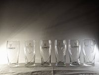 Front view of all six Guinness pint glasses lined up side by side showing various Guinness logos and harp icons in gold and black print.