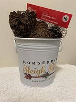 Front view of the white metal bucket with printed holiday text and a bag of cinnamon scented pine cones inside.