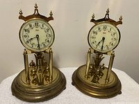 Two gold-tone Kundo anniversary clocks with white faces, side-by-side on a shelf against a wall