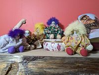 Five clown dolls arranged sitting on a rustic wooden surface with a reddish-orange wall background.