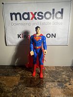 Front view of Superman action figure standing upright on a table with a maxsold banner in the background.