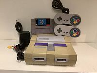 Front view of SNES console with Super Mario World cartridge inserted, two controllers, power adapter, and AV cable arranged behind.