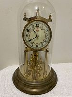 Full front view of the vintage clock under glass dome, showing brass base, columns, dial with floral pattern, and pendulum balls.