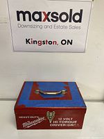 Red metal Milwaukee tool box with a silver handle on top, blue tape on lid, white text showing Heavy Duty Milwaukee 12 Volt Hi-Torque Driver/Drill.