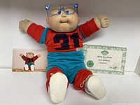 Photo of Cabbage Patch Kid Doll wearing blue glasses and red/blue outfit with birth certificate and a photo of the doll.