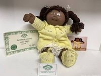 Full doll seated with certificate of authenticity and photo of the doll in the background.