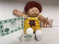 Cabbage Patch Kid doll seated showing yellow overalls with teddy bear design, brown curly hair, wearing white shoes, next to birth certificate and cards.