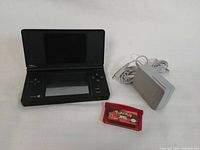 Black Nintendo DS Lite handheld console open with top screen and bottom touchscreen visible, placed next to grey power adapter and red translucent Pokemon Ruby game cartridge.