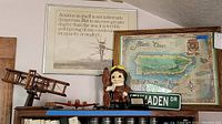 Photo showing wooden biplane model, framed aviation quote print, pilot figure, framed Puerto Rico map, and small road sign on top of bookshelf.