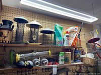 Shelf with bird feeders including metal mesh tube feeders, decorative rooster feeder, bird seed, gardening spray, and baskets on pegboard