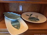 Two ceramic platters with fish design and one leaf-shaped bowl displayed on wooden shelving