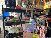 Wide shot of the black three-shelf metal wire cart with various items arranged on shelves and floor around it inside a garage or workshop.