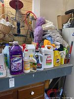 Photo showing cleaning liquid bottles, including Fabuloso, stain remover, dish soap, and bleach, alongside a stack of folded white towels and cleaning accessories on shelf.
