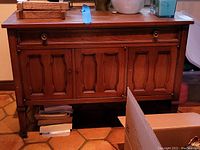 Front view of the wooden cabinet showing paneled doors, drawer, and two hardware pulls (one missing). The cabinet has a warm brown finish and shows scratches and wear.