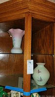 Photo showing two Fenton vases inside a wooden display cabinet: one with pink ruffled top and the other rounded with butterfly design.
